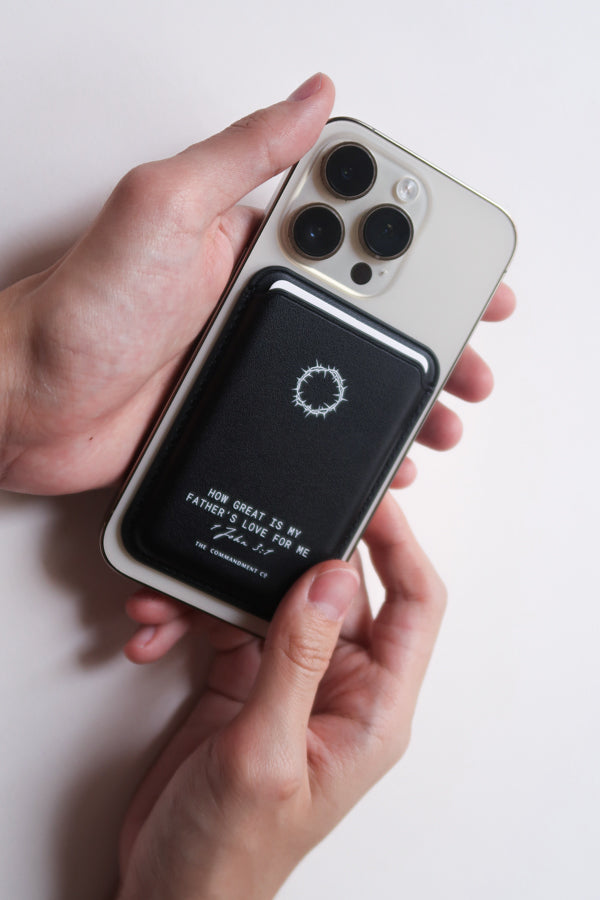 A pair of hands holding a mobile phone with the black Leather Magsafe Wallet attached to the back