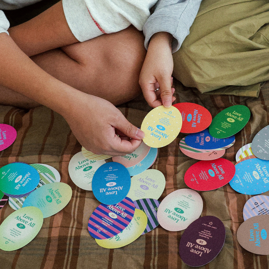 Hands picking a Love Above All card from a spread of colourful faith-based relationship cards on a bed during a quiet devotional moment.