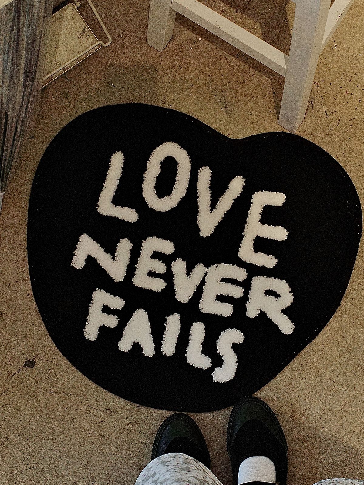 Black heart-shaped tufted rug with “Love Never Fails” lettering styled as Christian home decor indoors.
