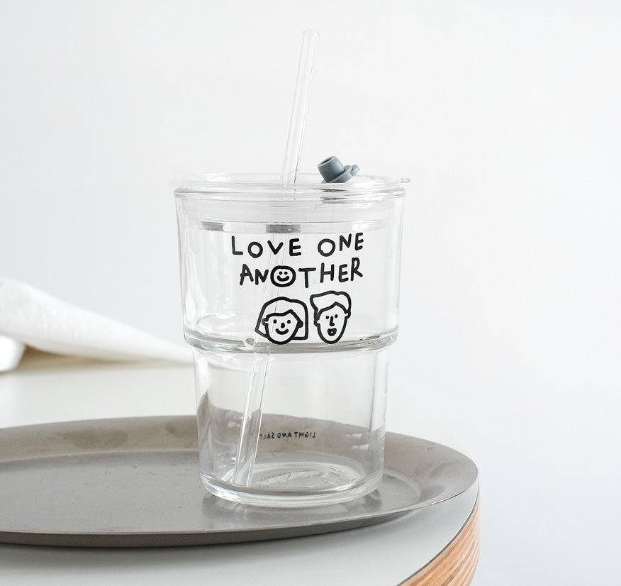 Clear glass tumbler with the phrase 'Love One Another' and cute hand-drawn faces, featuring a reusable straw, perfect for eco-friendly and inspirational drinkware.