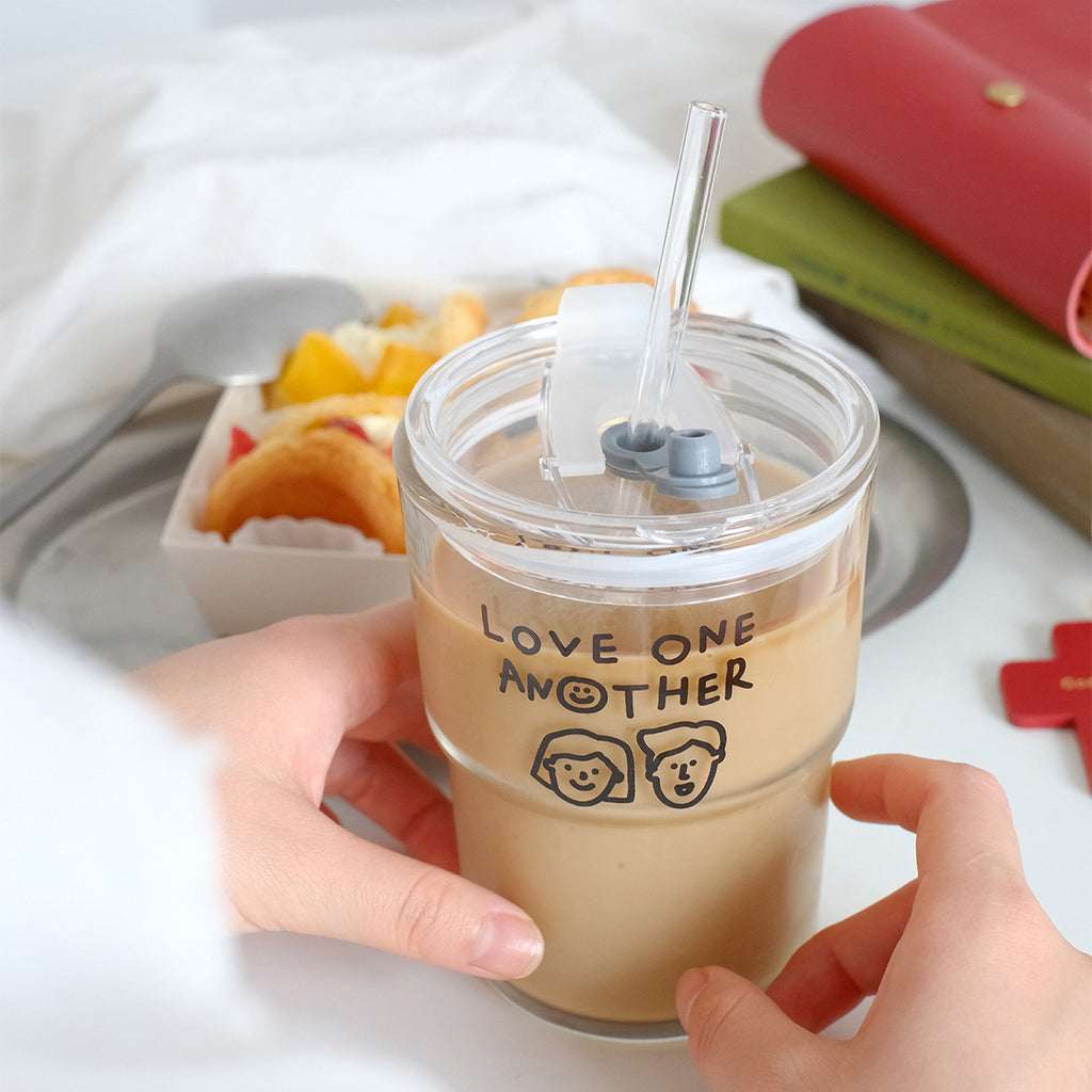 Hands holding a clear glass tumbler with 'Love One Another' design, filled with coffee, with a dessert box, books, and a red wallet in the background.