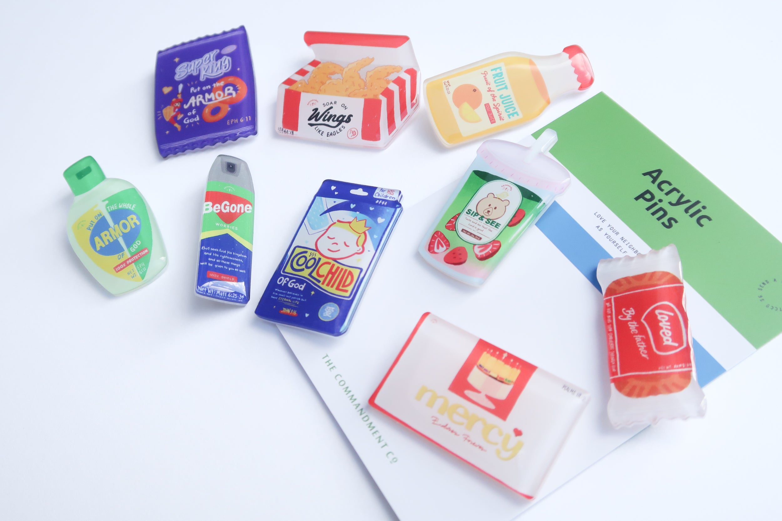 Full flatlay of Love Supermarket acrylic pin collection with Christian snack-themed designs, displayed alongside branded backing cards from The Commandment Co.