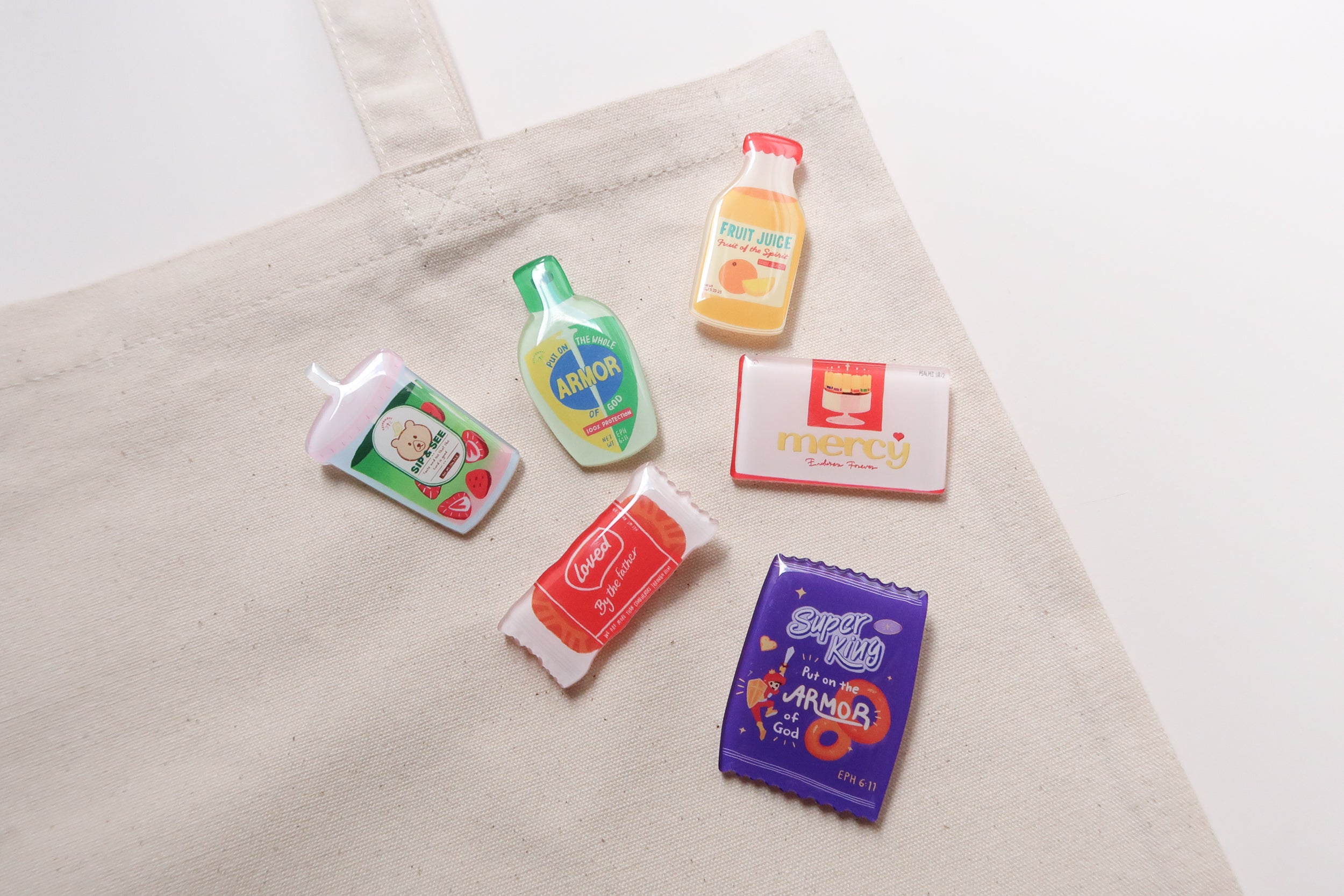 Close-up of Love Supermarket acrylic pins on a canvas tote bag, highlighting faith-based designs such as Armor of God, Fruit of the Spirit juice, Mercy chocolate, and Super King snack pin.