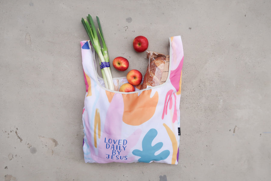 Foldable reusable 
shopping bag with Bible 
verse "Loved Daily by 
Jesus" in pastel colors, 
filled with groceries, a 
meaningful Christian gift 
from a Christian gift store. 