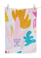 Colorful abstract tea towel with pink, yellow, and blue shapes and the phrase "LOVED DAILY BY JESUS" in blue; a Christian home decor gift from The Commandment Co.