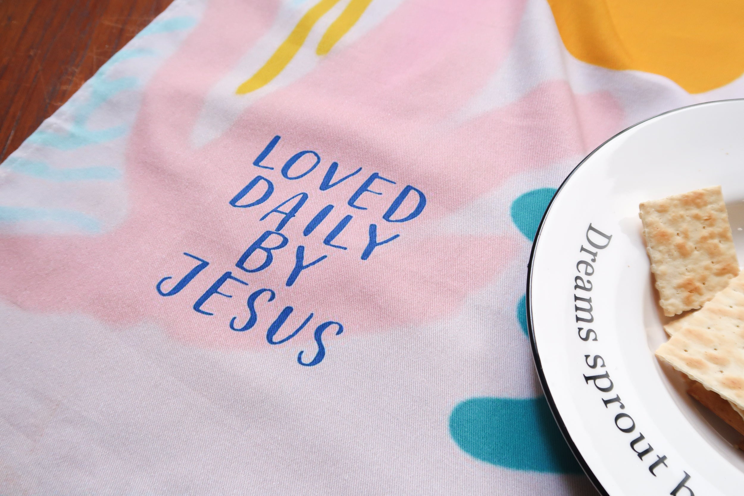 Pastel abstract tea towel with pink, yellow, and blue shapes and the blue text "LOVED DAILY BY JESUS"—a Christian home decor gift from The Commandment Co.