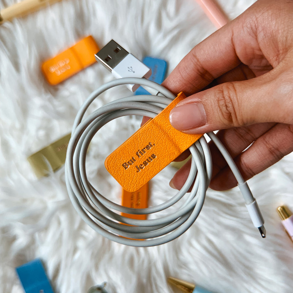 Orange magnetic bookmark labeled 'But first, Jesus' used to organize a USB cable, held by a hand over a white fluffy background with scattered bookmarks.