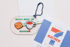  Acrylic keychain with "Match Made in Heaven" design featuring a biscuit and a cup of tea, alongside a Matthew 7:11 card, from The Commandment Co, Singapore.