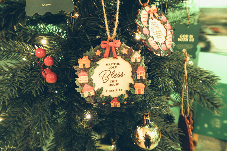 'May the Lord Bless This House' Christmas wreath ornament featuring small houses, holly leaves, and a red bow, hanging beautifully on a festive tree.