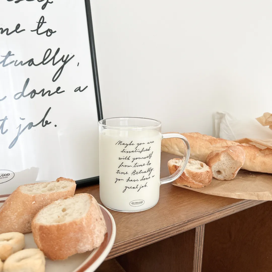 Clear glass mug with the encouraging message "Maybe you are dissatisfied with yourself from time to time. Actually, you have done a great job," filled with milk and surrounded by fresh bread, inspiring self-love and simple happiness.