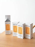 Three white milk cup cartons with an orange label and bottle illustration showing 350ml, arranged on a wooden table.