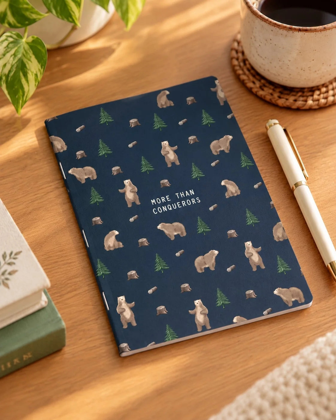 More Than Conquerors bear-pattern A6 notebook on a timber desk beside a pen and coffee.