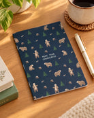 More Than Conquerors bear-pattern A6 notebook on a timber desk beside a pen and coffee.
