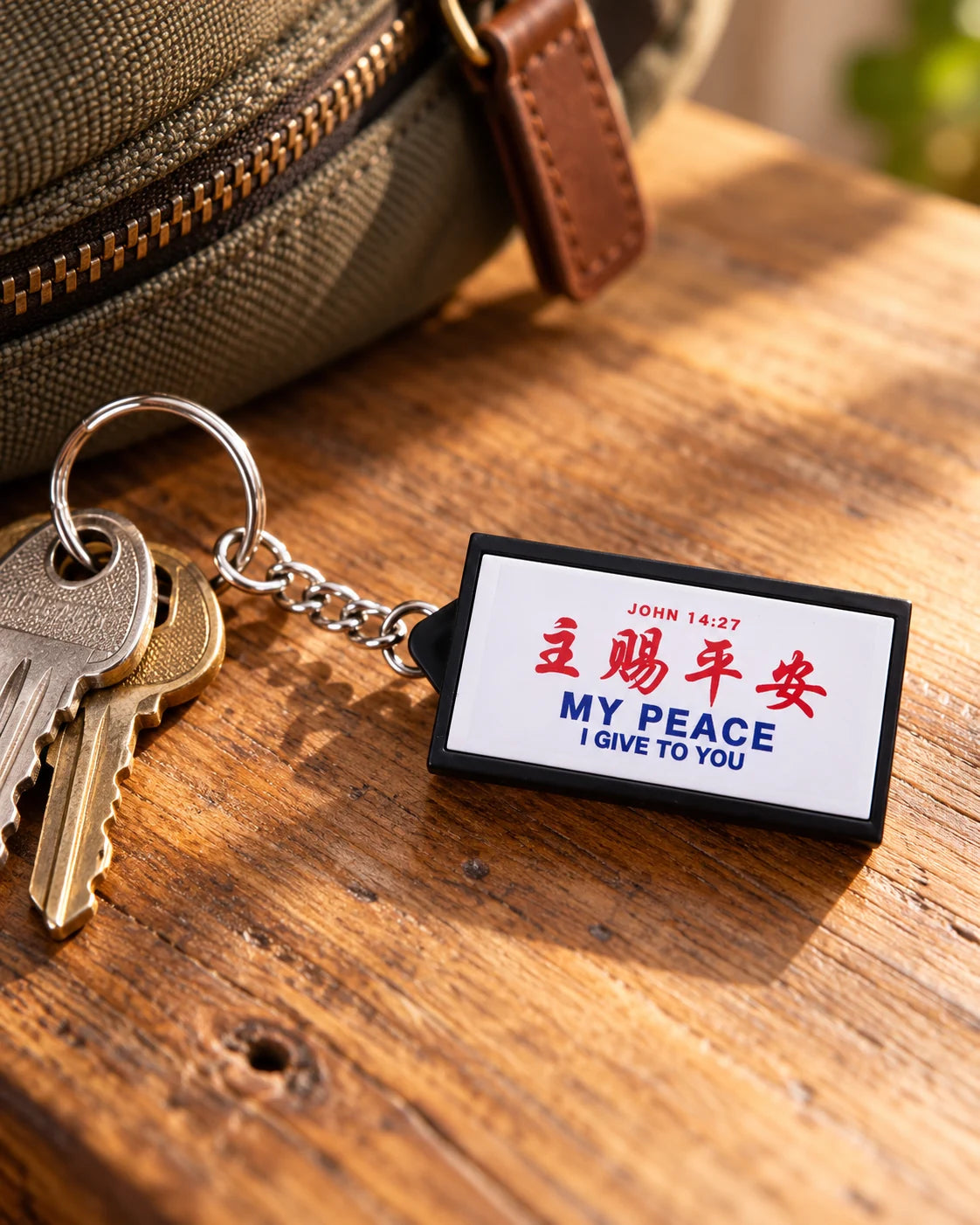 My Peace I Give To You bilingual lightbox keychain next to keys and a canvas bag on a wooden table.