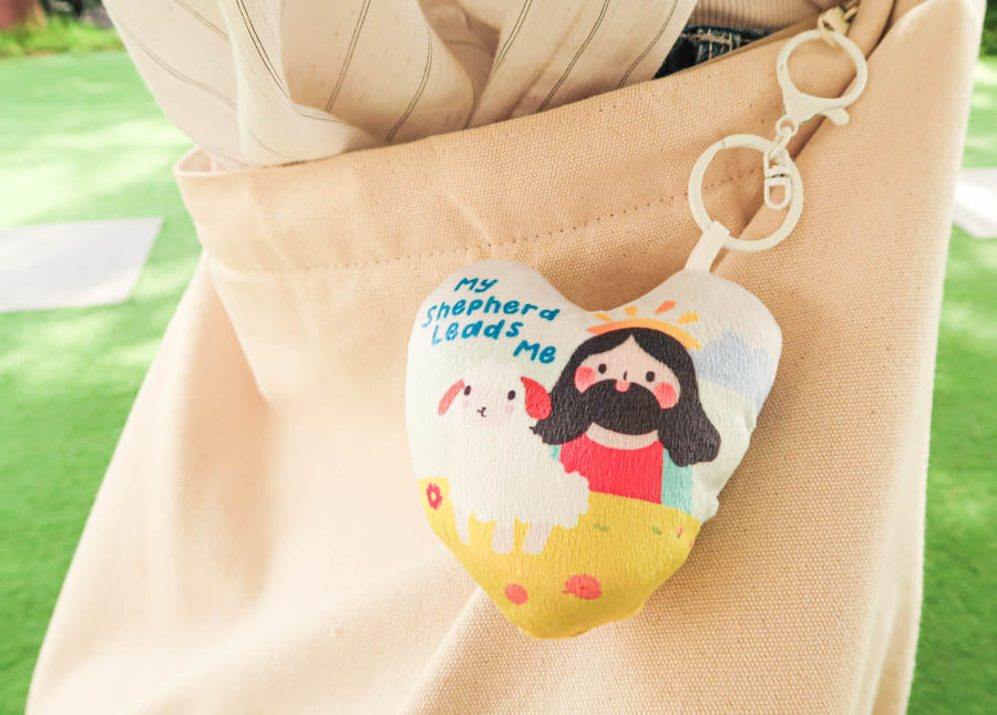 Heart-shaped plush keychain with 'My Shepherd Leads Me' text and illustration of Jesus with a sheep, attached to a beige bag.