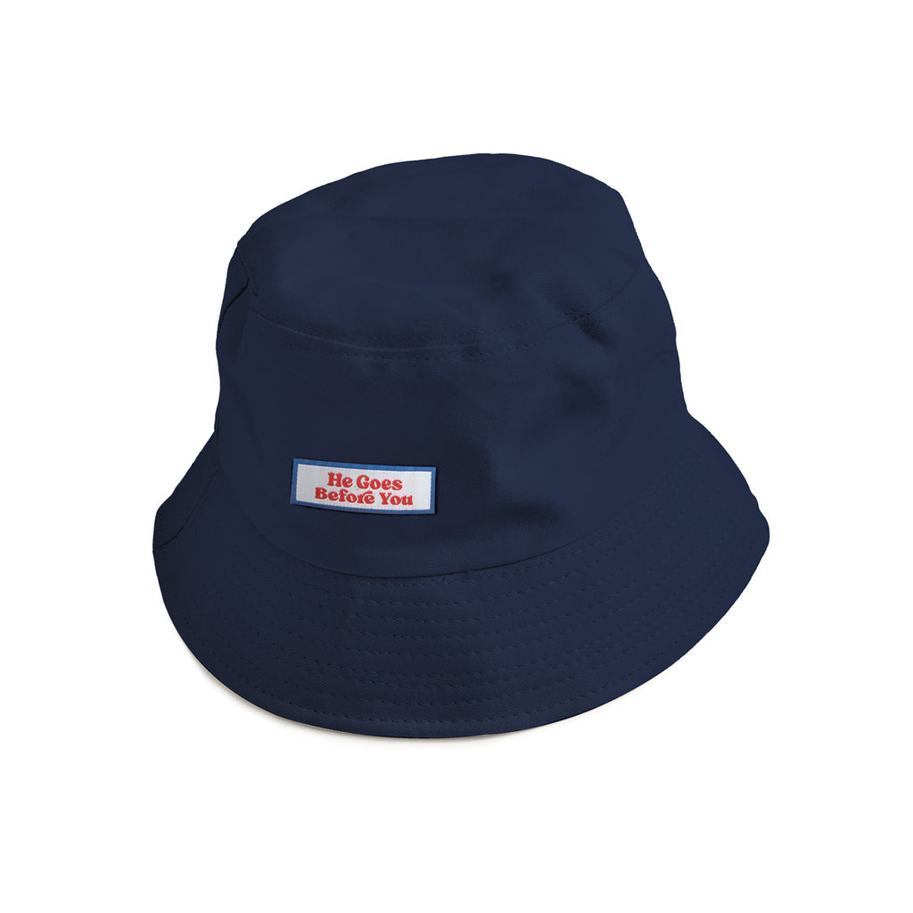 A navy blue bucket hat featuring a patch with red text reading "He Goes Before You," a faith-inspired Christian accessory for stylish outdoor wear.