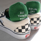 Green knit socks with white embroidery reading “ALL THINGS NEW” and “OLD LIFE HAS GONE,” nestled in a white checkered sneaker—a faith-filled daily reminder of renewal.
