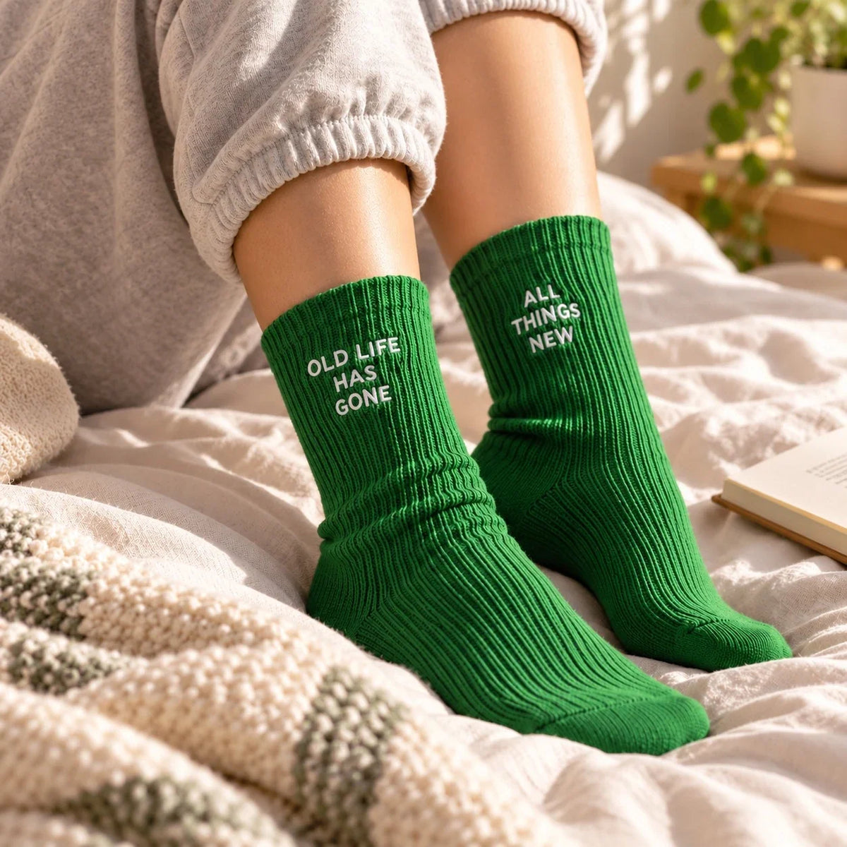Green Christian socks embroidered Old Life Has Gone and All Things New worn while relaxing on a cream bed.