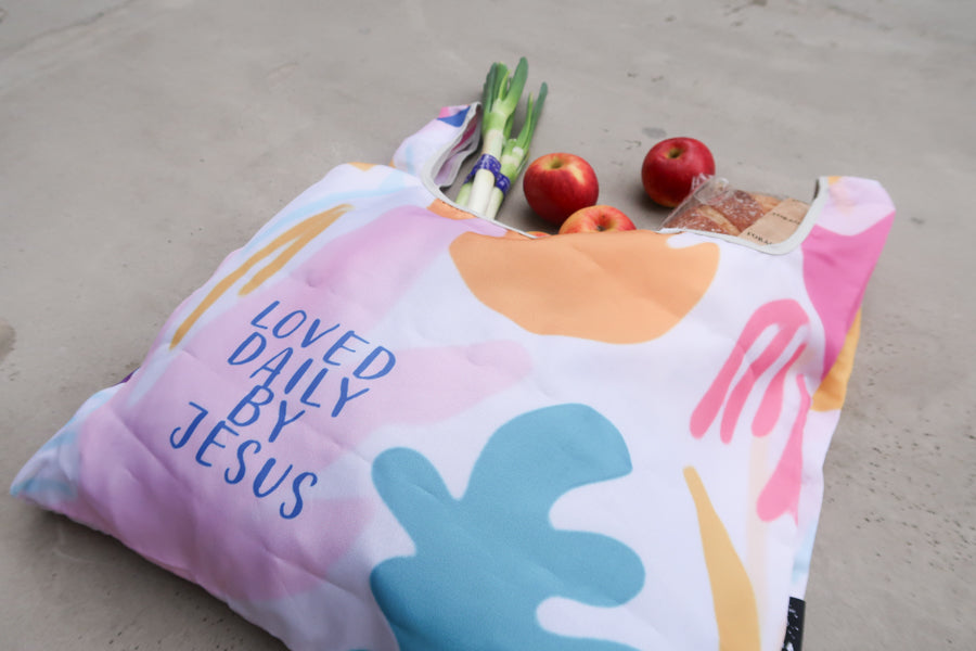 Close-up of pastel foldable 
reusable shopping bag with 
Bible verse "Loved Daily by 
Jesus," filled with 
groceries, a lovely Christian 
gift from a Christian gift 
store.