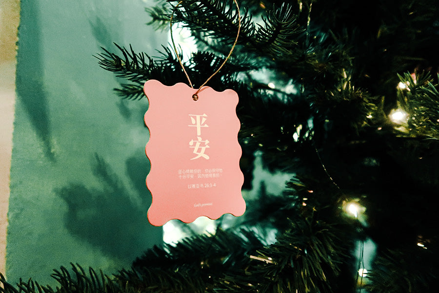 Christian Christmas tree hanging ornament featuring the Bible verse 'Peace' from Isaiah 26:3-4, with a pink scalloped design and twine string on a decorated tree.