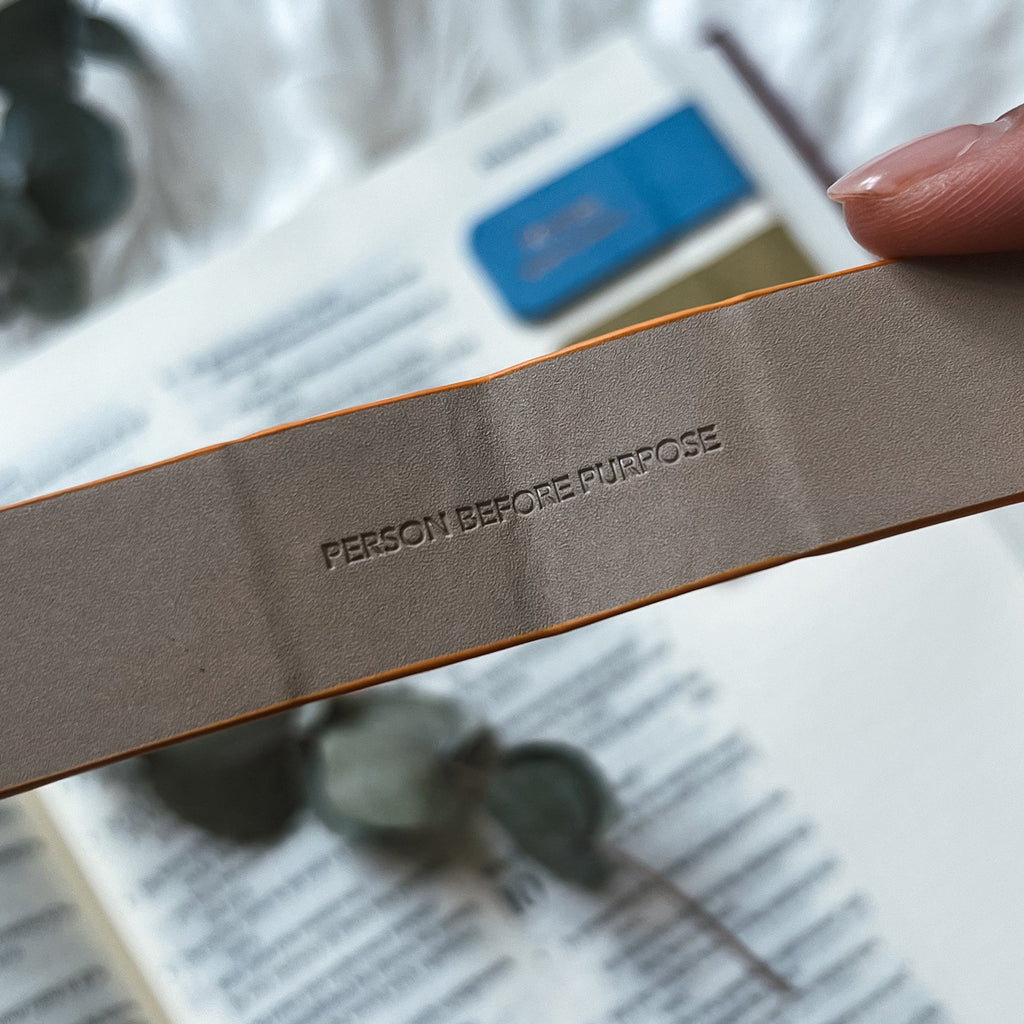 Close-up of a taupe-colored magnetic bookmark embossed with 'Person Before Purpose,' held over an open Bible with a blurred blue bookmark in the background