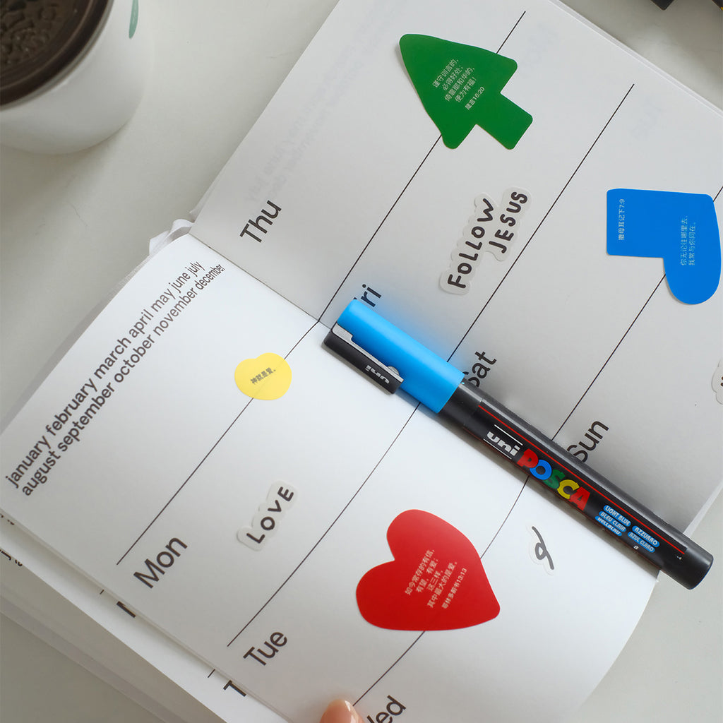 Open planner with colorful Bible verse stickers including a red heart, green tree, and text 'Follow Jesus,' along with a blue marker for faith-based decoration.