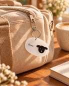 Pocket Psalms Grace sheep acrylic keychain clipped to the zipper of a beige travel bag in a warm sunlit setting.