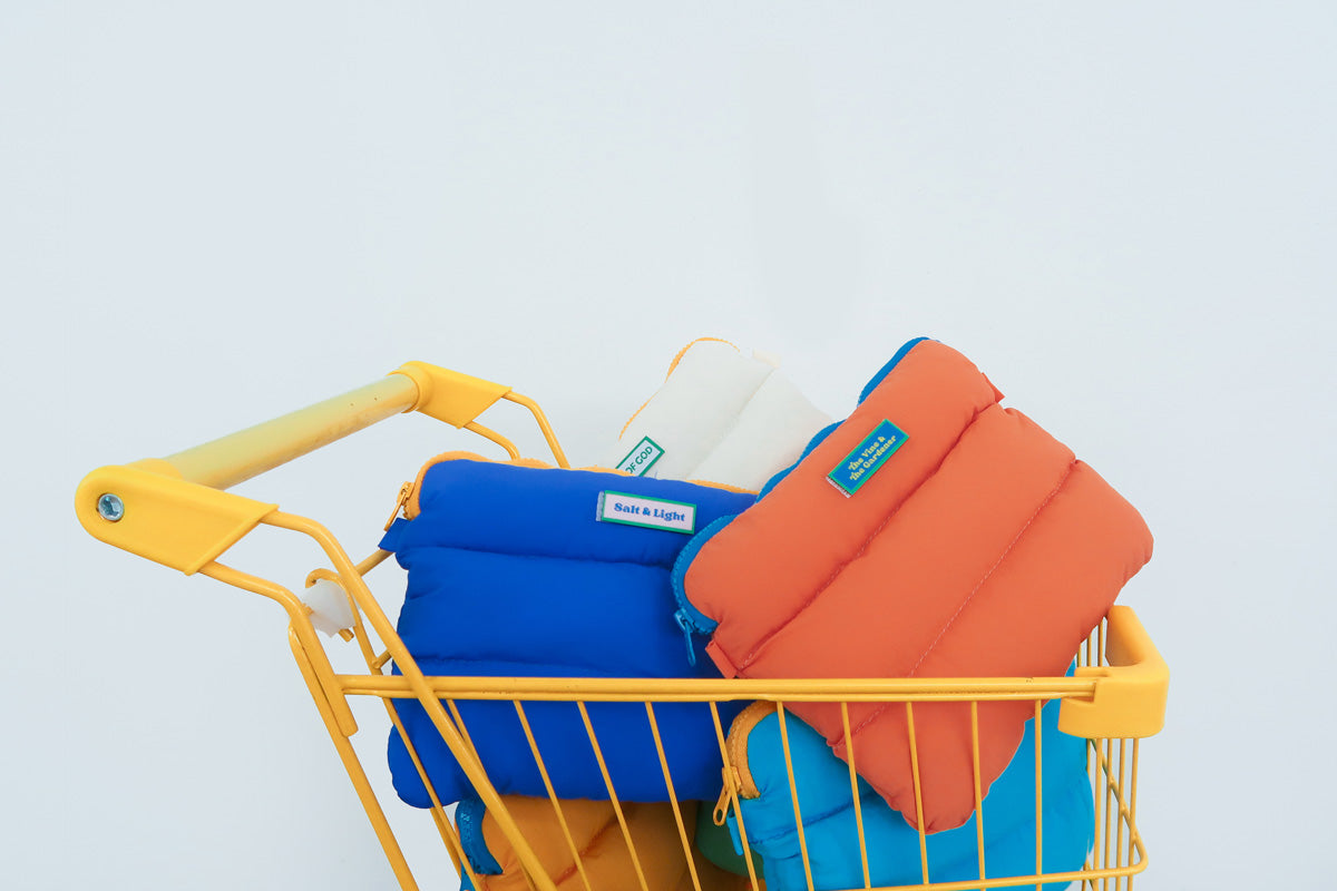 Colorful Pong Pong Bags displayed in a vibrant yellow shopping cart, featuring blue, orange, and white designs with unique velcro patches that read 'Salt & Light' and 'The Vine & The Gardener.'