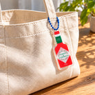 Power & Strength chili sauce acrylic keychain hanging from a cream canvas tote bag.