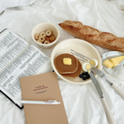Profite du moment présent A5 notebook with pen, open Bible, pancakes with butter, blueberries, boiled eggs, cereal, and a baguette on a cozy white bed, inspiring simple happiness and mindful living.