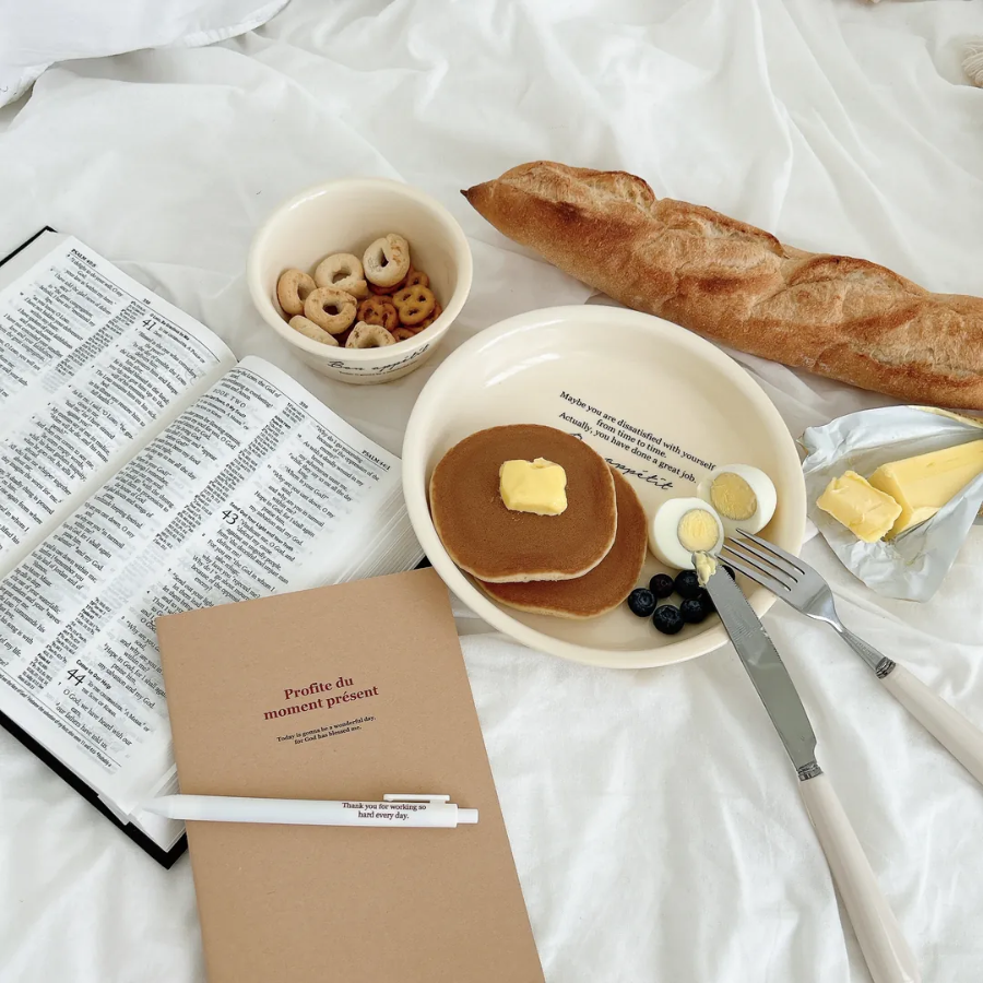 Profite du moment présent A5 notebook with pen, open Bible, pancakes with butter, blueberries, boiled eggs, cereal, and a baguette on a cozy white bed, inspiring simple happiness and mindful living.