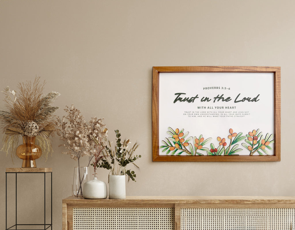 A wooden-framed wall art featuring the Bible verse "Trust in the Lord" from Proverbs 3:5-6. The artwork is decorated with vibrant orange and green botanical designs. It is displayed in a neutral-toned room with a rattan cabinet adorned with dried flowers in vases, creating a modern yet rustic and serene ambiance ideal for Christian home decor.