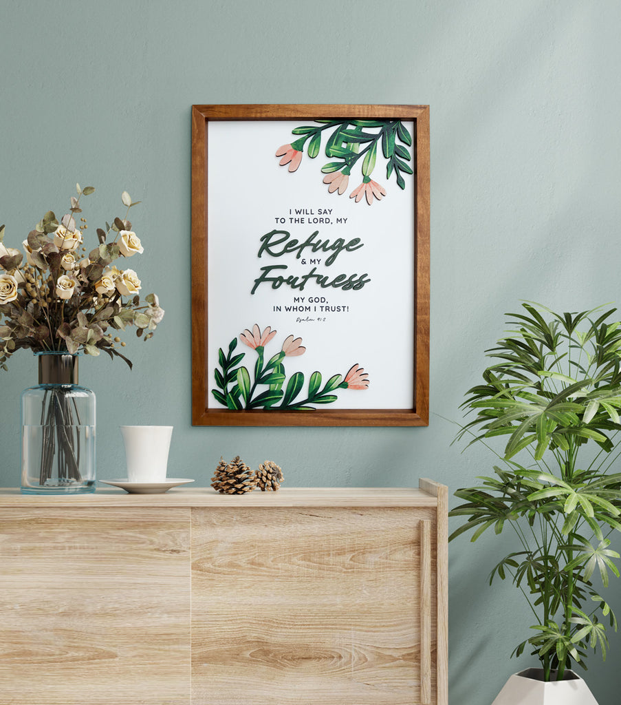 A beautifully designed wooden-framed wall art featuring the Bible verse "I will say to the Lord, my Refuge and my Fortress, my God, in whom I trust" from Psalm 91:2. The artwork is adorned with delicate green leaves and pink flowers, creating a serene and elegant appearance. Displayed on a wooden console with a clear glass vase holding dried flowers, a white ceramic cup, and decorative pinecones, complemented by a leafy green plant against a muted green wall.