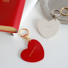 Two heart-shaped keychains displayed next to a red book: a red keychain with 'Love above all' and a white keychain with 'Hallelujah,' featuring gold and silver rings.