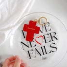 Red cross-shaped keychain engraved with 'God is love,' placed on a glass plate with 'Love Never Fails' text, a Christian faith-inspired accessory.