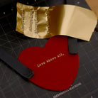 Red heart-shaped keychain with 'Love above all' embossed in gold foil, displayed alongside an embossing machine and gold foil material.