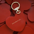 Red heart-shaped keychain with 'Love above all' engraved in gold, placed on a background of red heart cutouts, representing love and faith.