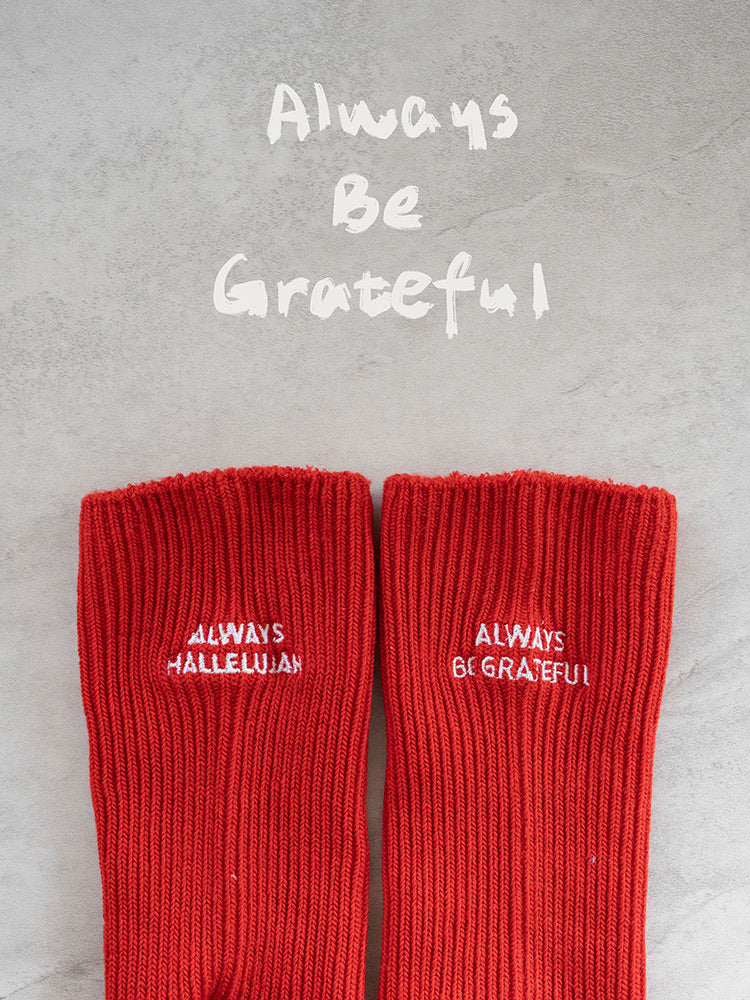Red knit socks with white embroidery reading “Always Hallelujah” and “Always Be Grateful” on a gray backdrop, a daily Christian faith reminder.