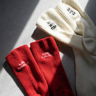 Red ribbed socks with white embroidery reading “Always Hallelujah” and “Always Be Grateful” placed beside cream knit socks; a daily faith reminder.