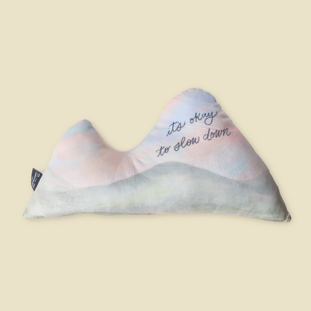 Soft, mountain-shaped plush pillow in calming pastel colors with the gentle reminder 'it's okay to slow down,' perfect for Christian home decor and relaxation.