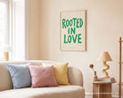Rooted In Love Christian poster displayed above a cream sofa in a warm living room with pastel cushions and a table lamp
