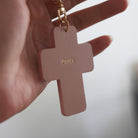 Rose pink cross-shaped keychain with 'Peace' engraved, held in a hand, symbolizing faith and serenity.