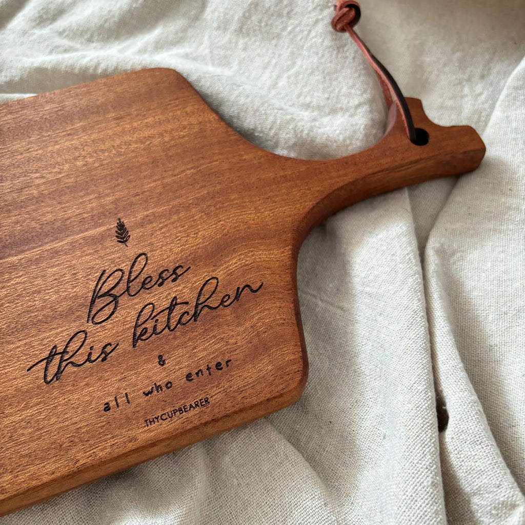 A rustic wooden cheese board engraved with the phrase "Bless this kitchen and all who enter," accompanied by a wheat symbol. The board features a leather strap for hanging and is styled on a soft beige fabric, making it a charming addition to kitchen decor or a thoughtful gift.