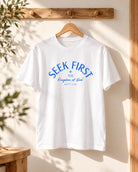 Seek First Christian white T-shirt with Matthew 6:33 hanging on a wooden hanger in a sunlit room.