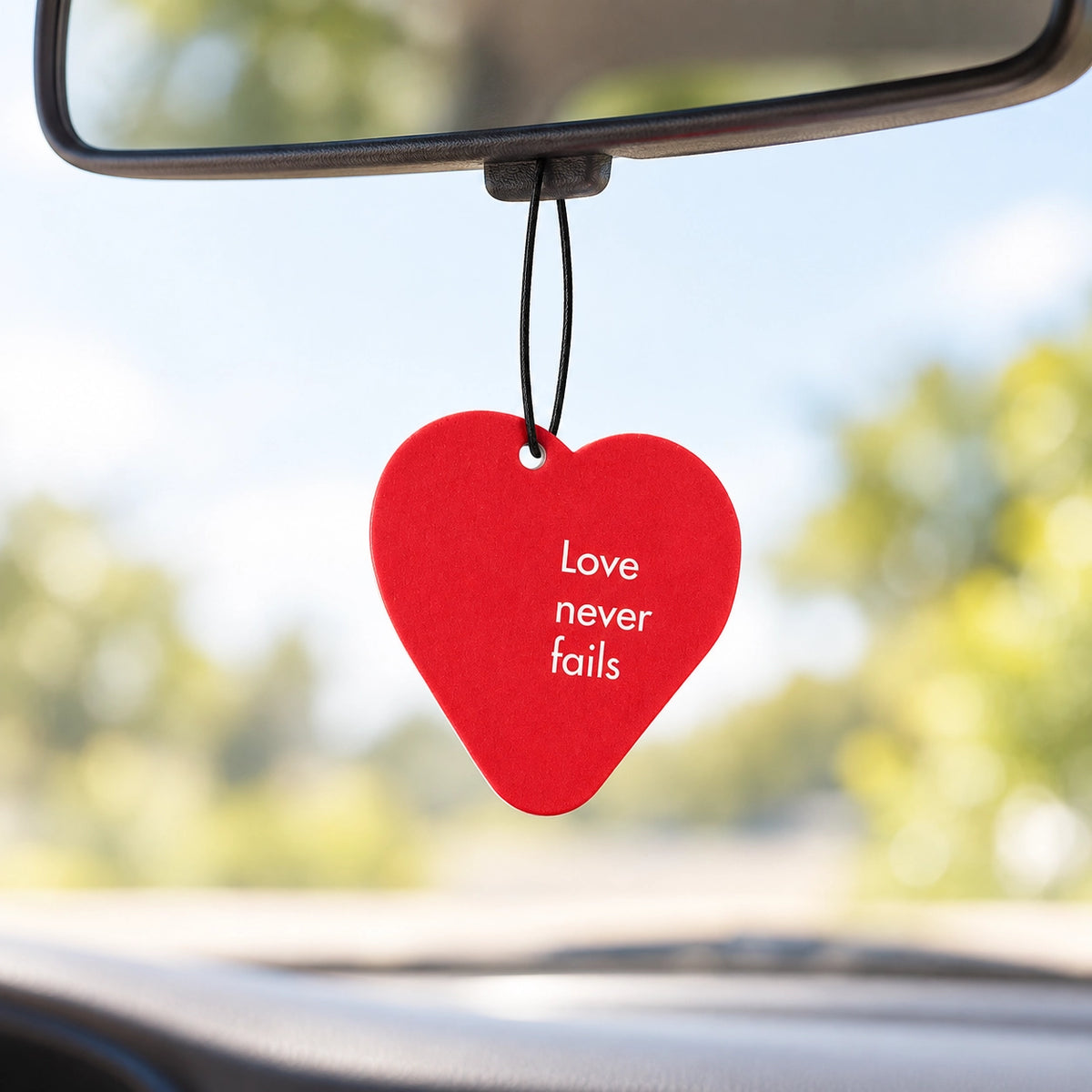 Love Never Fails red heart scent tag hanging from a car rear-view mirror