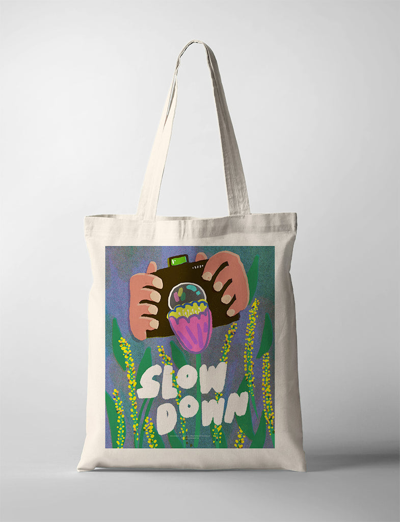 Inspirational tote bag with 'Slow Down' text and colorful illustration of hands holding a camera