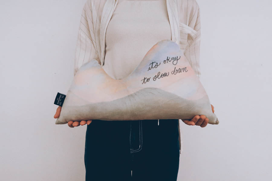 Person holding a mountain-shaped plush cushion with the message 'it's okay to slow down,' designed to inspire relaxation and mindfulness.