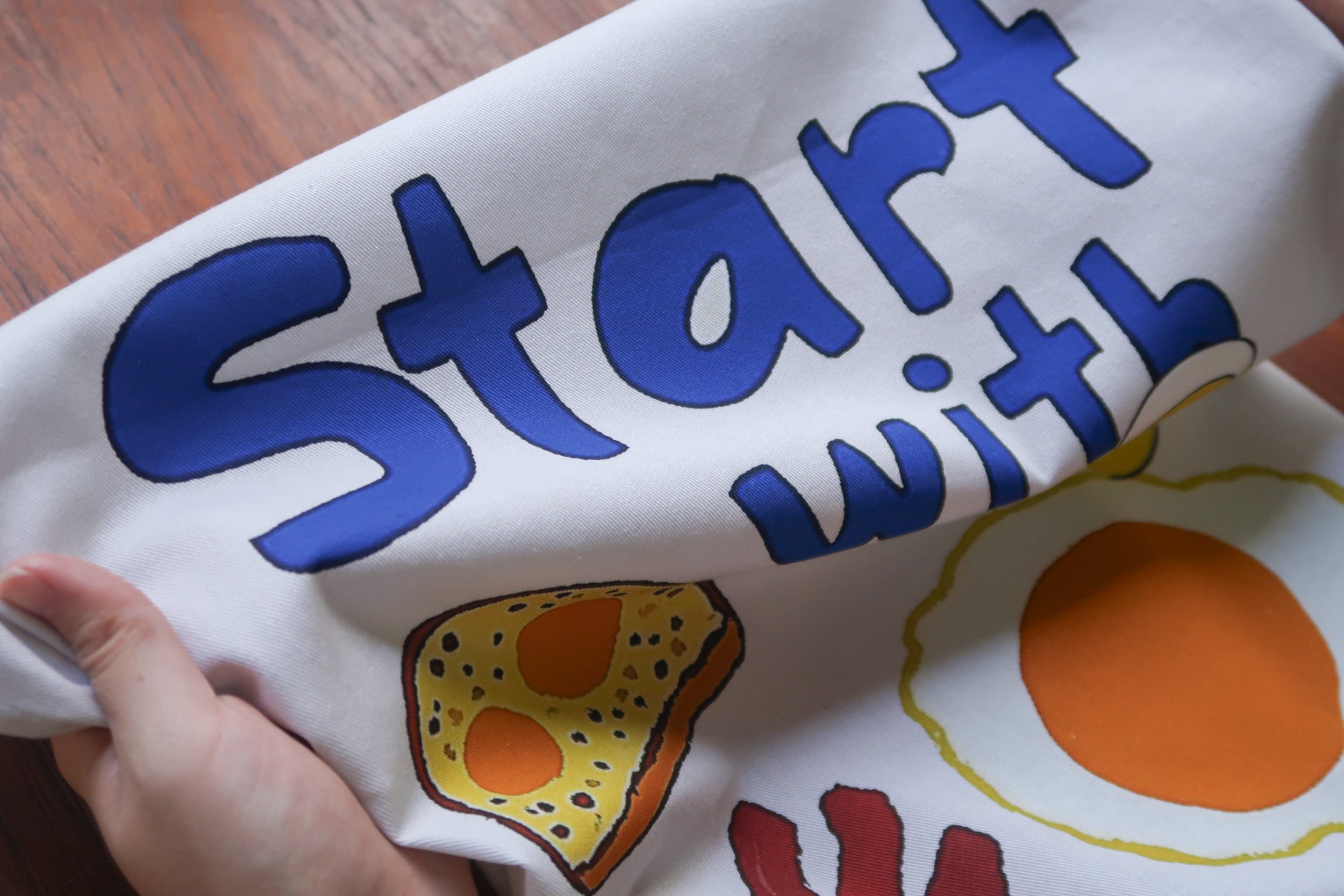 Start With Jesus tea towel with bold blue typography and breakfast illustrations (toast and fried egg) on a white cotton towel—a contemporary Christian home decor gift.