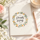 Strength & Dignity A6 Christian notebook styled with flowers, coffee and a pink pen.