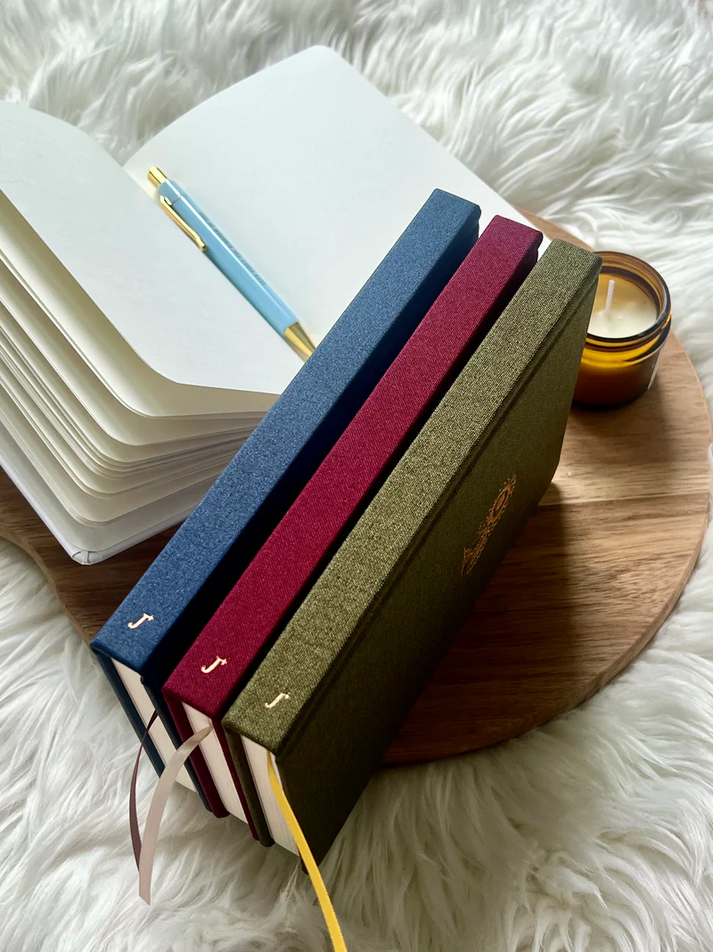 Three fabric hardcover journals by Project J in blue, red, and green displayed beside an open notebook and candle on a wooden tray.